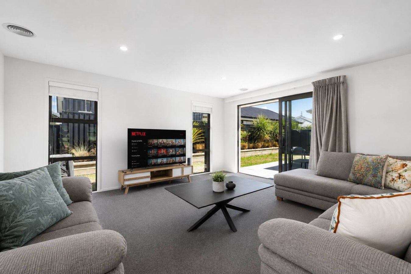 A modern living room with a flat-screen TV, two sofas, a coffee table, and a sliding glass door leading to a garden.