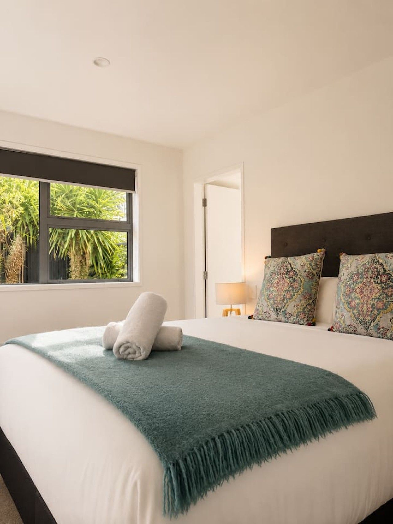 A bedroom with a bed, a window, a door, and a nightstand with a lamp.