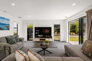 A modern living room with a flat-screen TV, two sofas, a coffee table, and a large window with a view of greenery. - Mountain View - 2 Bedroom House (Queenstown)