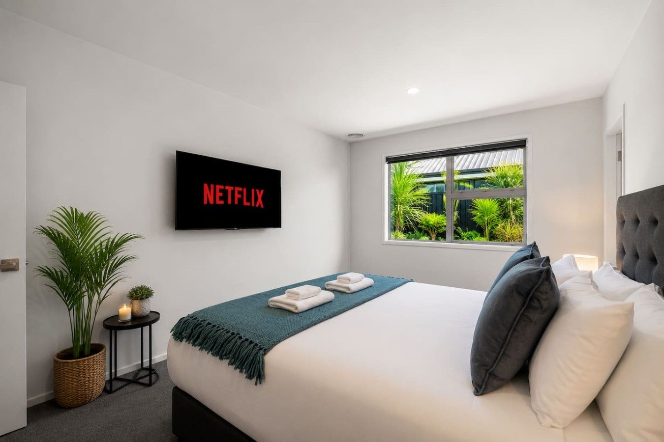 Habitación de hotel moderna con cama, televisor mostrando 'NETFLIX', ventana con vista a follaje y planta en maceta.
