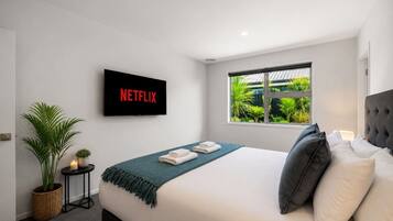 Habitación de hotel moderna con cama, televisor mostrando 'NETFLIX', ventana con vista a follaje y planta en maceta.