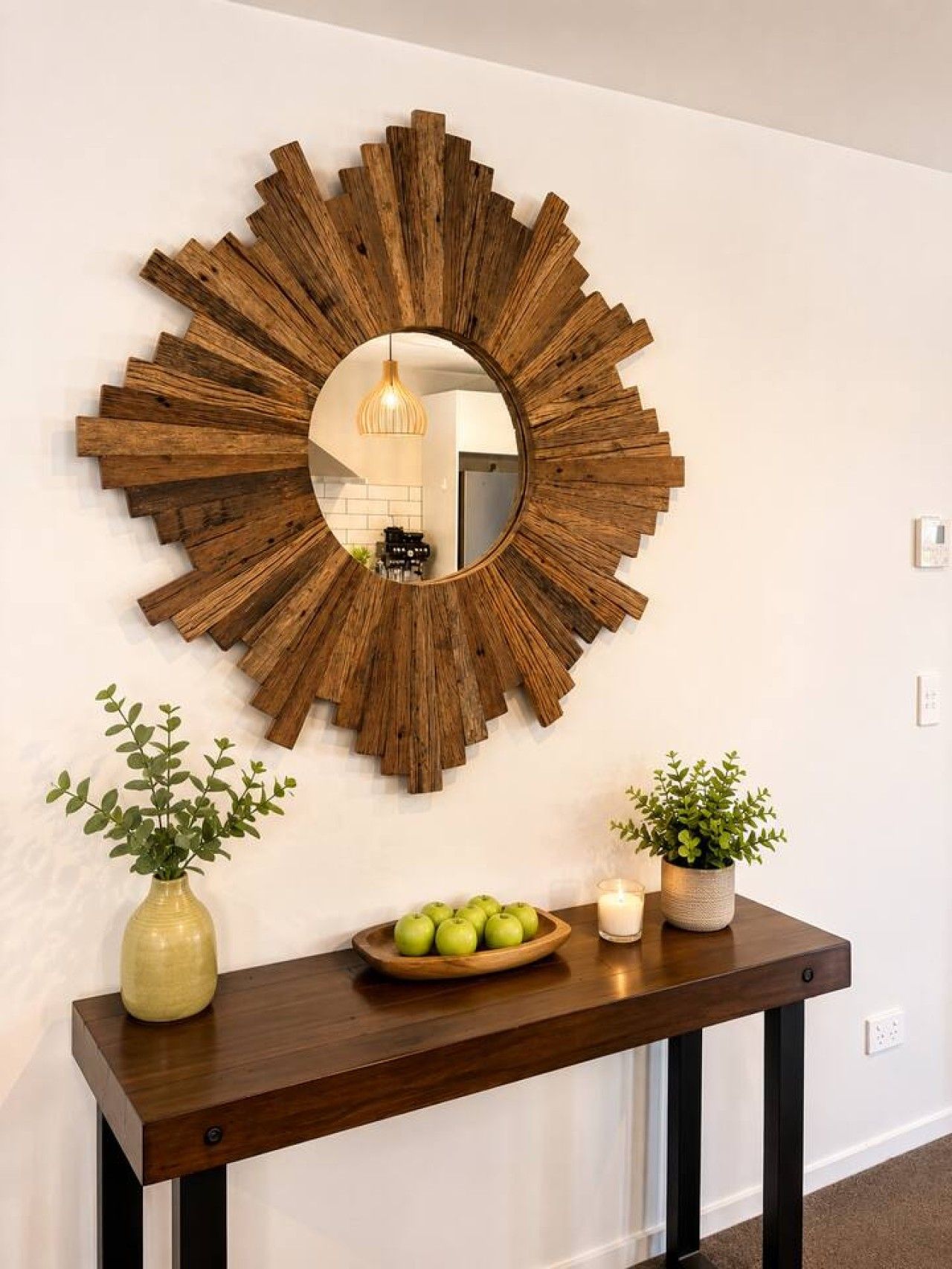 A wall-mounted wooden sunburst mirror with a round mirror in the center, a wooden console table with a bowl of green apples, two potted plants, and a lit candle.