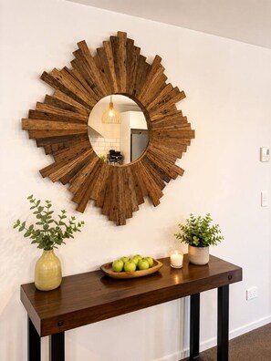 A wall-mounted wooden sunburst mirror with a round mirror in the center, a wooden console table with a bowl of green apples, two potted plants, and a lit candle.