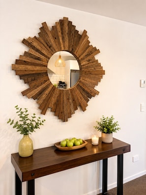 A wooden wall decor with a mirror, a wooden table with a bowl of apples, and two potted plants.