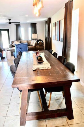 A dining area with a long wooden table, chairs, a small decorative item, and a view of a living room with a sofa and curtains. - Wine Country Retreat | Saint Jo 3BR Home + Pool & Hot Tub (Saint Jo)
