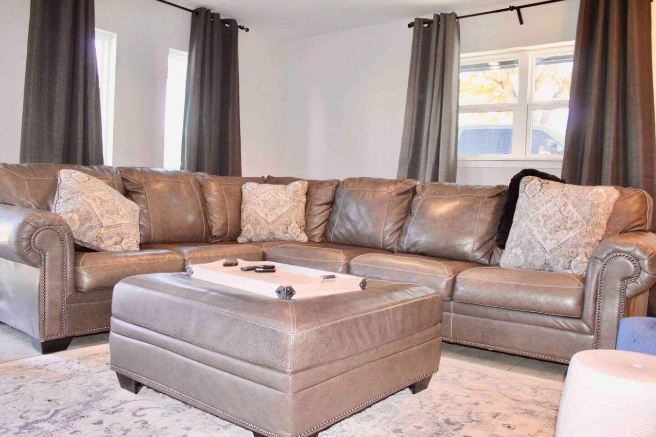 A sectional sofa with a matching ottoman in a living room with a large window and curtains.