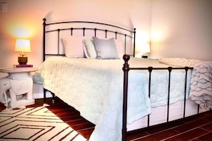 A neatly made bed with a white duvet and pillows, a bedside table with a lamp, and a patterned rug on a wooden floor. - Wine Country Retreat | Saint Jo 3BR Home + Pool & Hot Tub (Saint Jo)