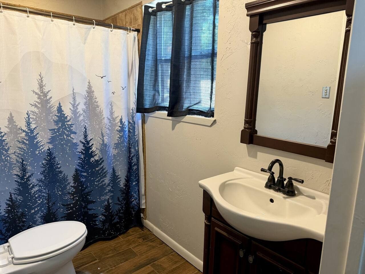 Une salle de bain comprenant des toilettes, un lavabo avec un miroir et un rideau de douche orné d’un motif de forêt.