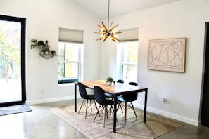 A dining area with a wooden table, black chairs, a woven rug, a pendant light, and a wall-mounted abstract art piece. - Stylish Nocona Barndo | Wineries + Fire Pit (Nocona)