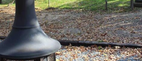 A black outdoor heater with a chimney, surrounded by gravel and fallen leaves, with a house and trees in the background.