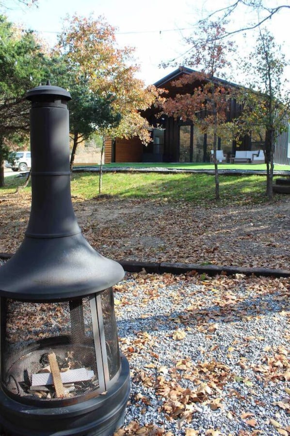 Un calentador exterior negro con una chimenea, rodeado de grava y hojas secas, con una casa y árboles al fondo.