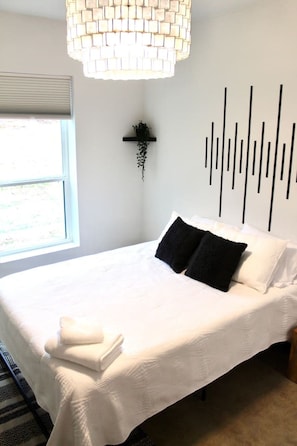 A bedroom with a large bed, a wall-mounted plant, a black vertical light fixture, and a window with blinds. - Stylish Nocona Barndo | Wineries + Fire Pit (Nocona)