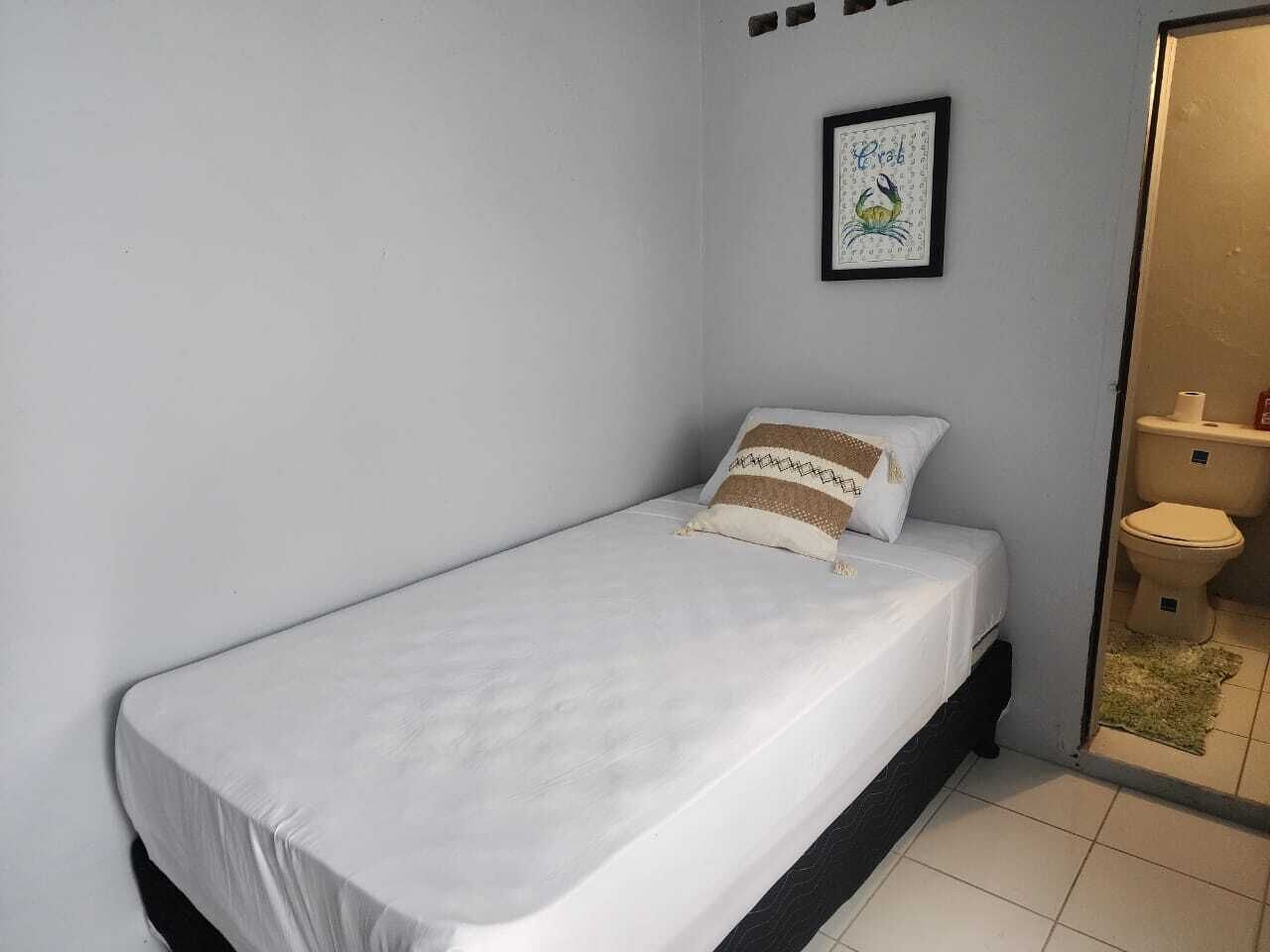 Una habitación pequeña con cama individual, colcha blanca, almohada estampada y una obra de arte enmarcada en la pared.