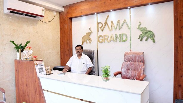 Una reception con un uomo seduto dietro, un pannello di legno con 'RAMA GRAND' e sagome di animali, una pianta in vaso e una sedia di plastica.