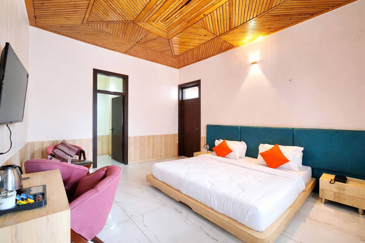 A hotel room with a wooden ceiling, a bed with white bedding and orange pillows, a pink chair, a wooden nightstand, and a television.