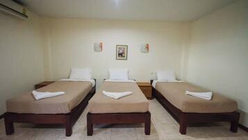 Habitación de hotel con tres camas individuales, cada una con una almohada blanca y una toalla doblada. El piso es de baldosas y la pared blanca, sin decorar, tiene un cuadro enmarcado sobre las camas.
