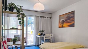 A hotel room with a bed, a desk with a chair, a window with curtains, a plant on the desk, and a painting on the wall.