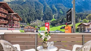 A balcony with white plastic chairs and a small table with a vase of flowers and two wine glasses. In the background, there is a mountain landscape with a ski resort and a sign for 'On the Rocks'.