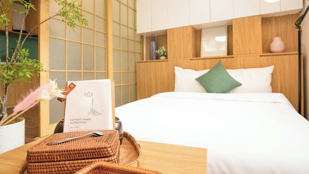 A hotel room with a bed, a bedside table with a lamp, a vase with flowers, and a wooden wall with a sliding door. - The Setai Apartments Linh Lang by XÔI Residences (Hanoi)