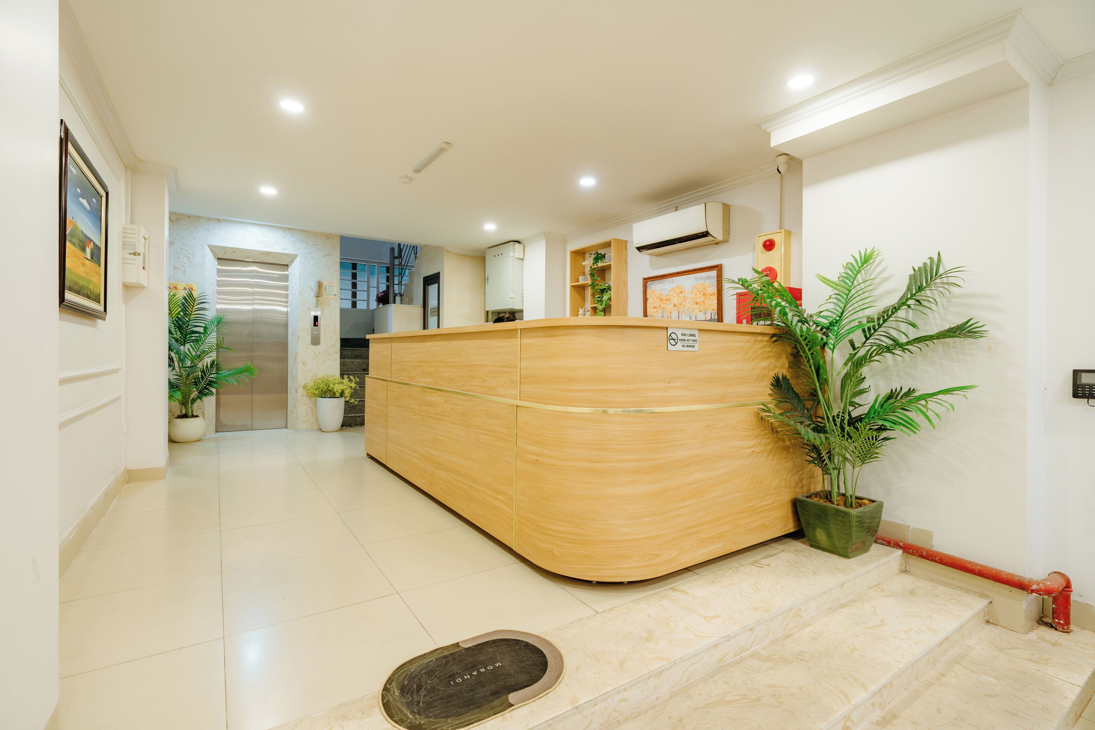 Un área de recepción con un mostrador de madera, plantas en macetas, una barandilla y un ascensor visible.