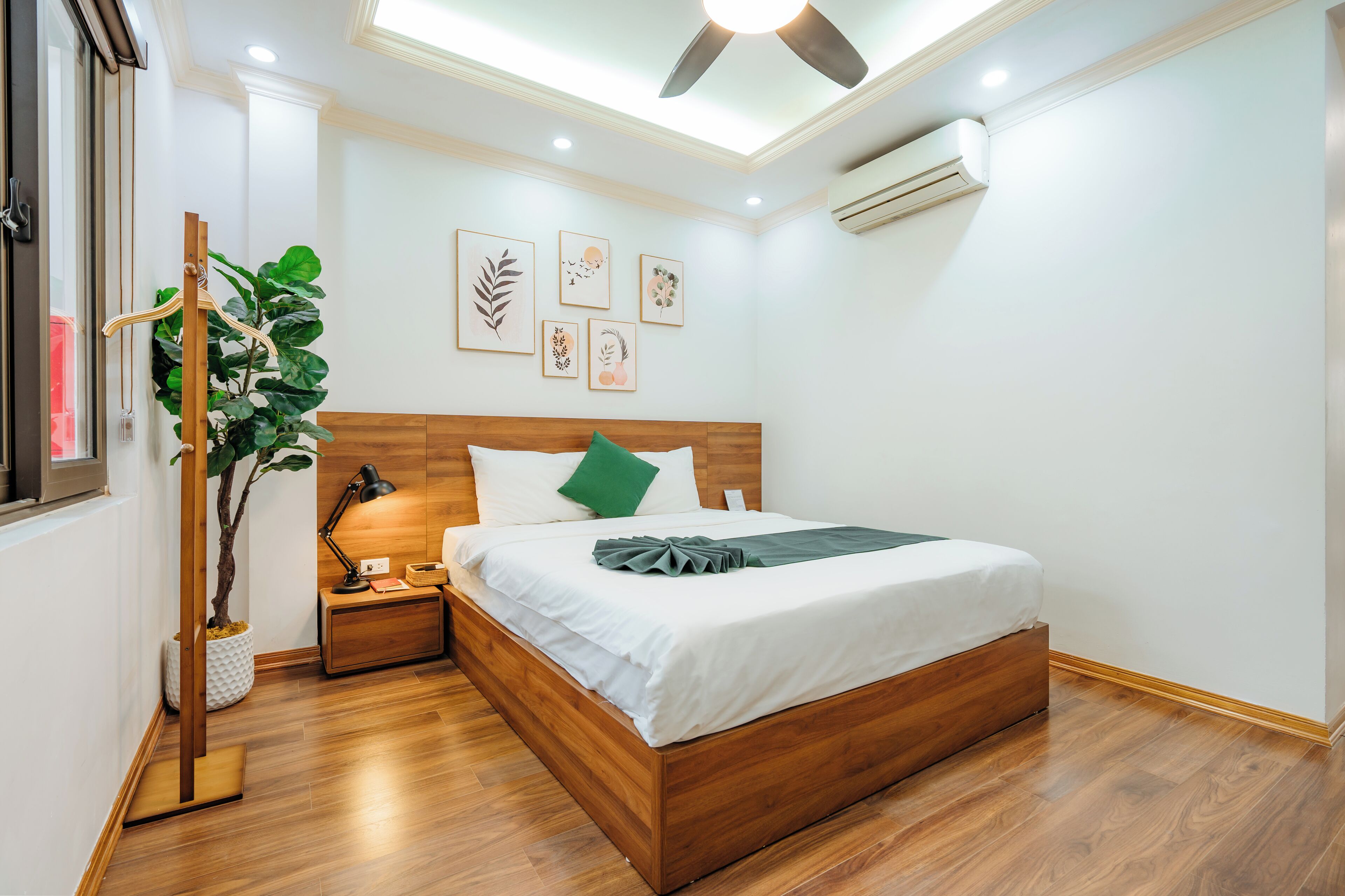 A neatly arranged bedroom with a wooden bed, a bedside table with a lamp, a wall-mounted air conditioner, and framed artwork.