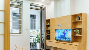 A modern hotel room with a wooden TV stand, a flat-screen TV displaying a VR app, a small table with a vase and a basket, and a balcony with a potted plant. - The Setai Apartments Linh Lang by XÔI Residences (Hanoi)