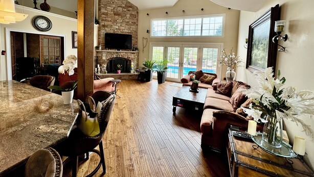 A spacious living room with a stone fireplace, a large TV, and a view of a swimming pool. - Luxury 5BR Family Getaway – Heated Pool, Hot Tub, Fire pit, Game Room, (Roselle)