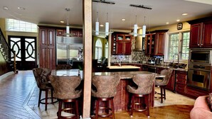 A spacious kitchen with a central island, wooden bar stools, stainless steel appliances, and a herringbone tile backsplash. - Luxury 5BR Family Getaway – Heated Pool, Hot Tub, Fire pit, Game Room, (Roselle)