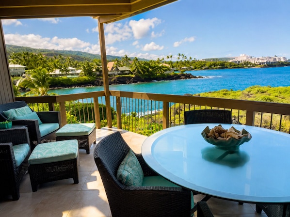 A covered outdoor seating area with wicker furniture and a glass table, overlooking a coastal view with palm trees and a clear blue sky.