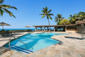 A pool area with lounge chairs and umbrellas, a covered seating area, and a view of the ocean.