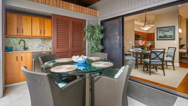 A dining area with a glass table surrounded by wicker chairs, a kitchen with wooden cabinets and a tiled backsplash, and a sliding glass door leading to another room.
