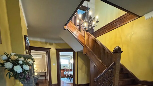 A wooden staircase with a chandelier, leading to an upper floor. A stone wall and a flower arrangement are visible on the left. - 6 BR, 4 BA Mansion Close to the City (Yonkers)