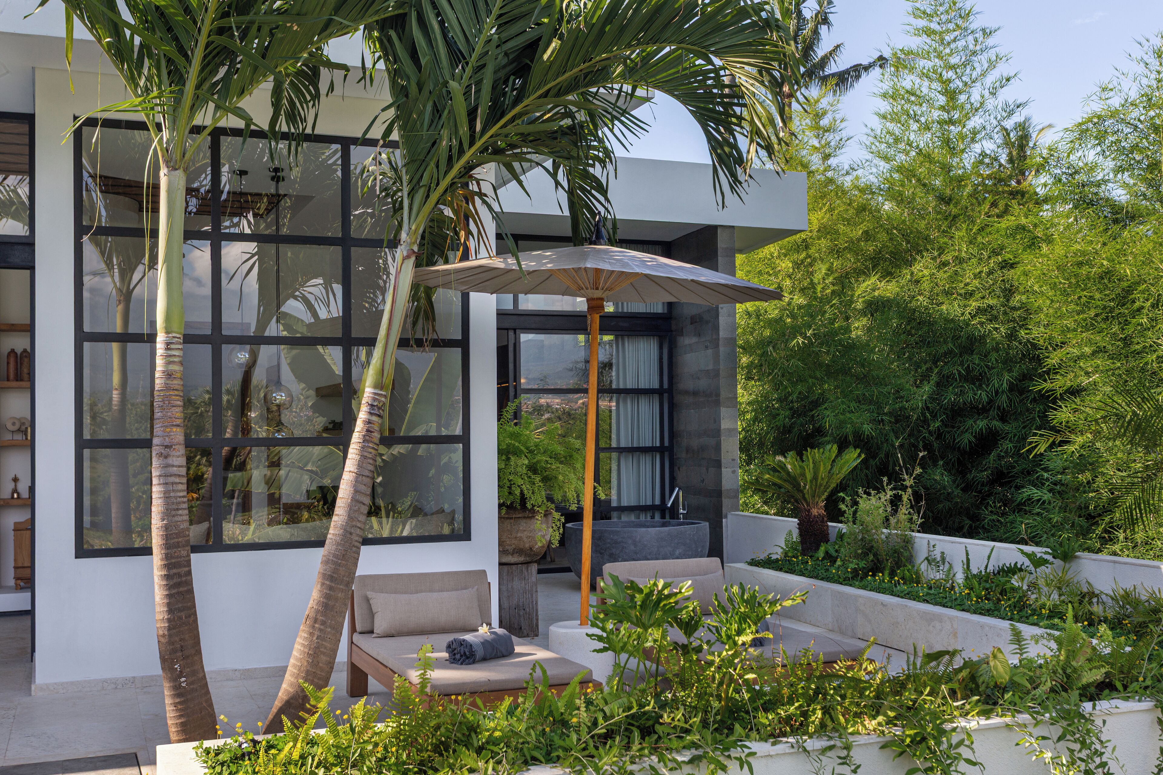 A modern outdoor seating area with a sofa and an umbrella, surrounded by lush greenery and palm trees.