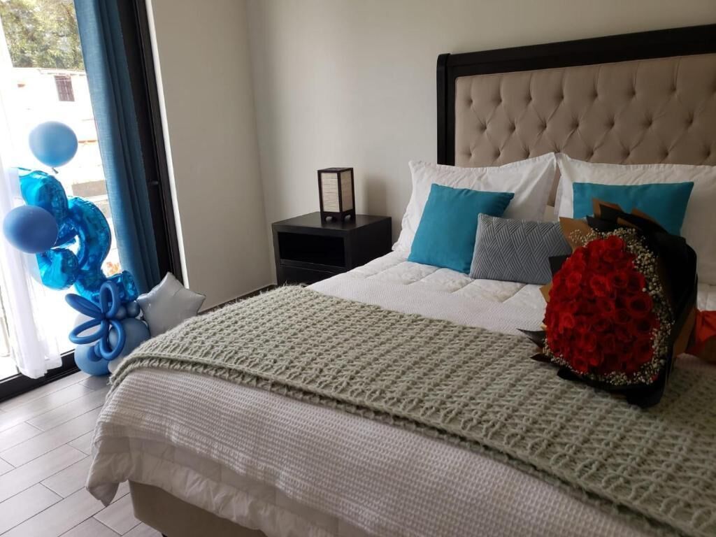 A neatly made bed with a white duvet, a grey pillow, and a red floral pillow. There is a black nightstand with a framed object on it.