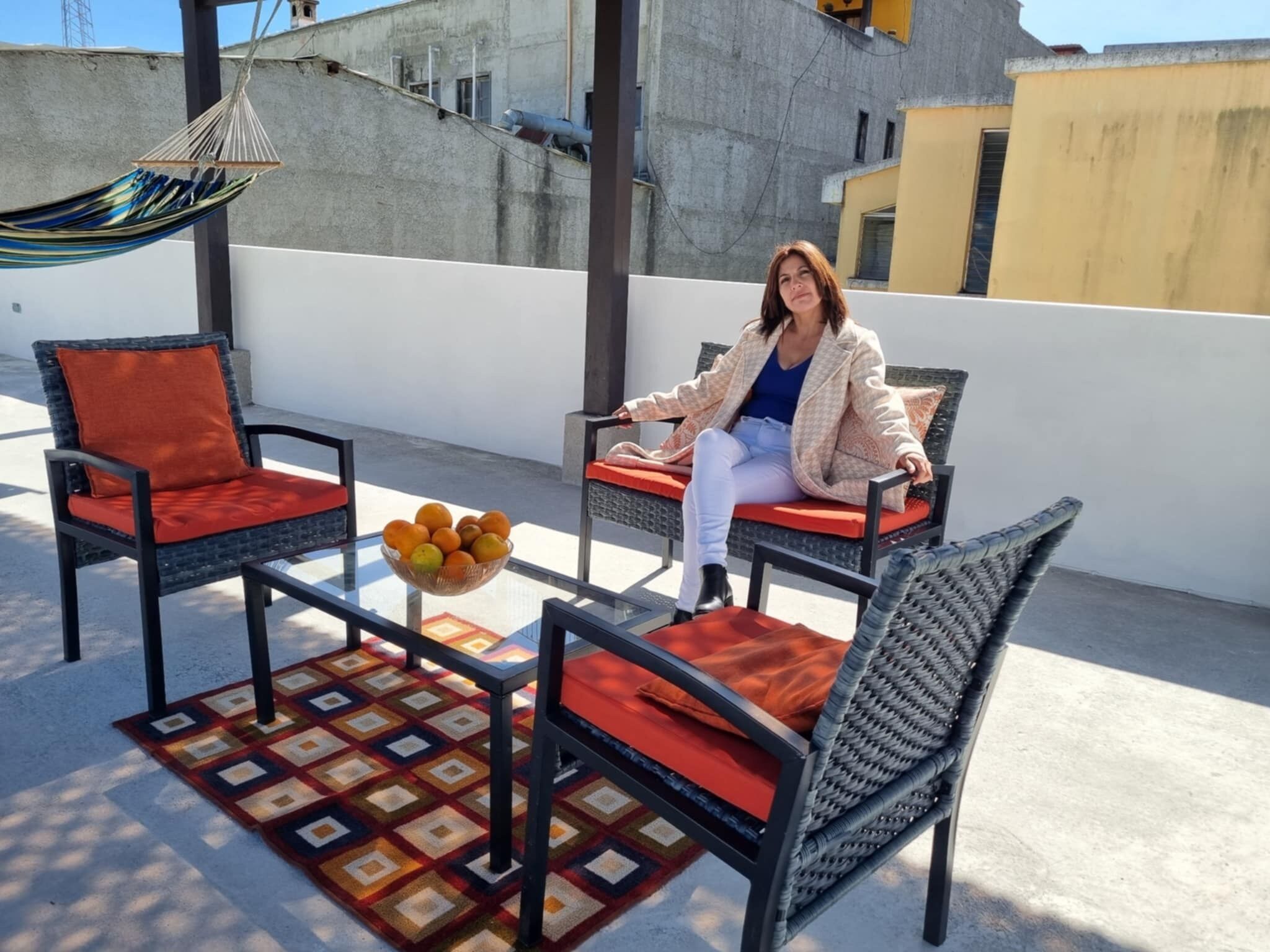 Un balcón con mobiliario de mimbre, una cesta de fruta tejida y un tapete estampado.