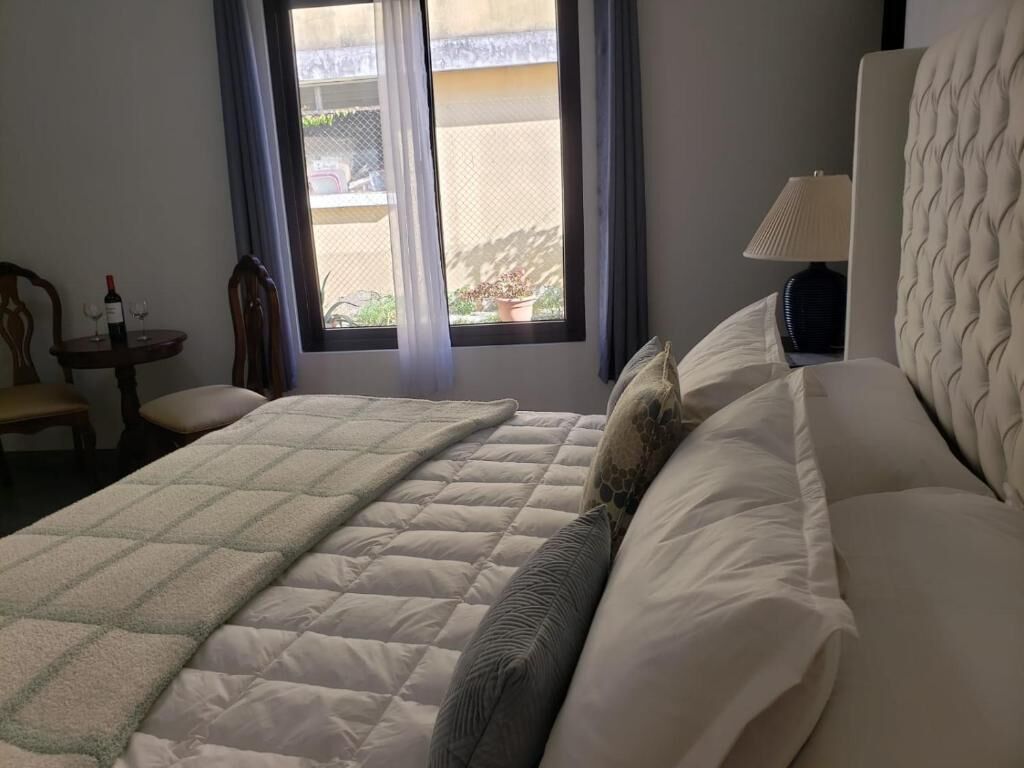 A hotel room with a bed, a bedside table with a bottle and glasses, a lamp, and a window with curtains.