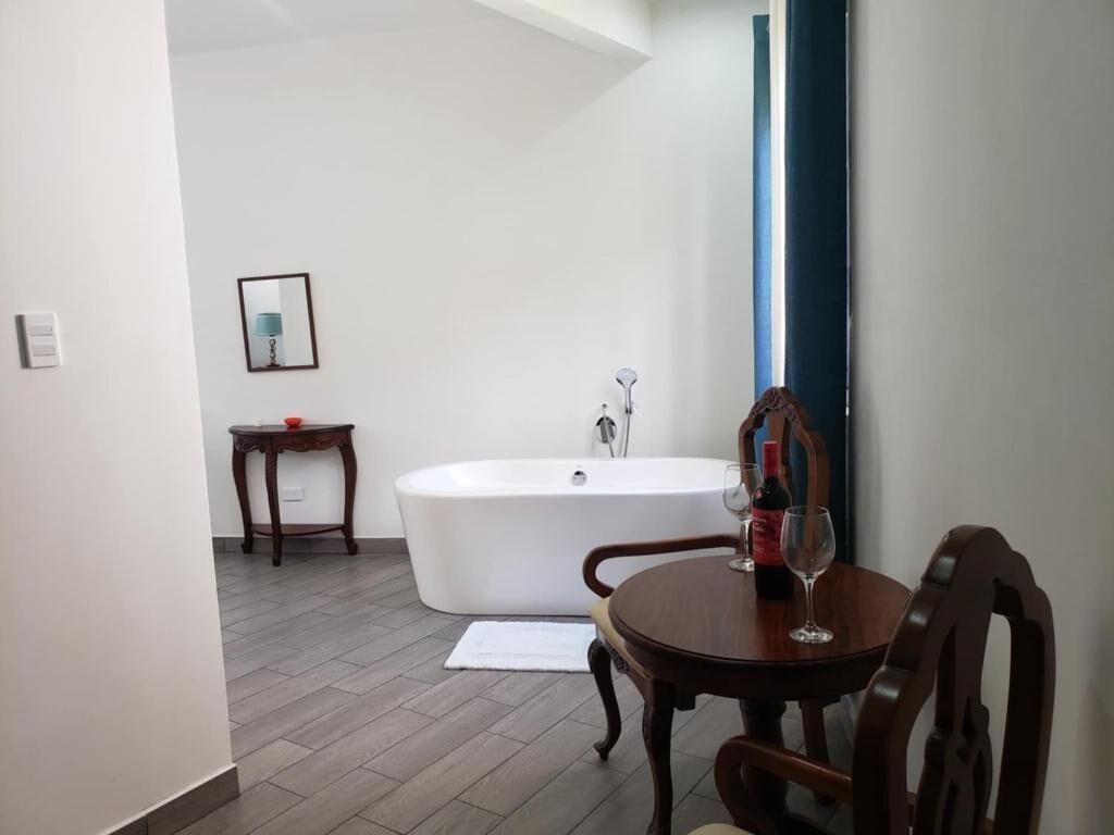 A bathroom with a bathtub, a small table with a bottle and glass, and a chair.