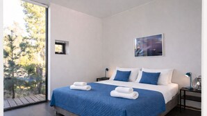 A modern bedroom with a large bed, two bedside tables, a window with a view of trees, and a painting on the wall.