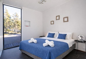 A modern hotel room with a large bed, a view of trees through a sliding door, and a small table with a fan.