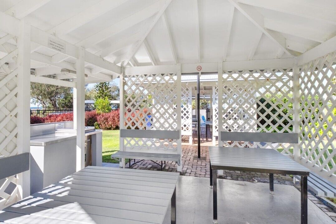 A covered outdoor seating area with benches and tables, surrounded by a white lattice fence.