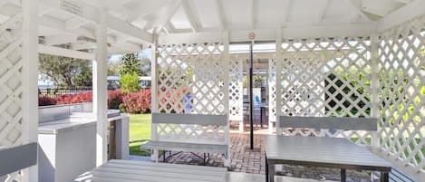 A covered outdoor seating area with benches and tables, surrounded by a white lattice fence.