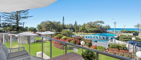 A balcony with a view of a pool, garden, and beach.