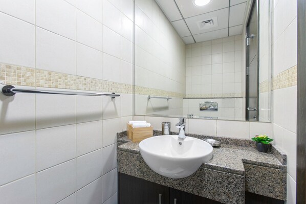 Un baño moderno con azulejos blancos, encimera de granito, lavabo blanco, espejo y zona de ducha.