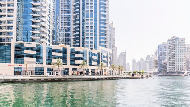 Een hotel aan het water met moderne architectuur, een promenade met palmbomen en uitzicht op het water.
