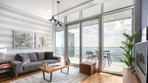 A modern living room with a grey sofa, a wooden coffee table, and a large window offering a city view.