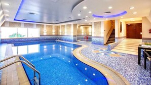 Une grande piscine intérieure à la forme incurvée, entourée d’un sol et de murs recouverts de carreaux, et un escalier menant à un niveau supérieur.