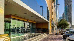 Un bâtiment moderne doté d’une façade en verre, d’une entrée pavée de briques et d’un espace de stationnement où sont garées des voitures.