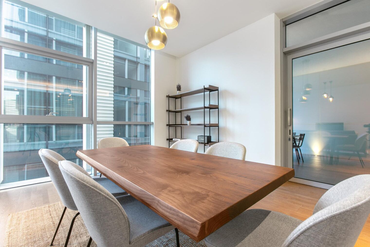 Una sala de reuniones moderna con una gran mesa de madera, sillas tapizadas en gris, un estante de metal negro y grandes ventanales con vistas a la ciudad.