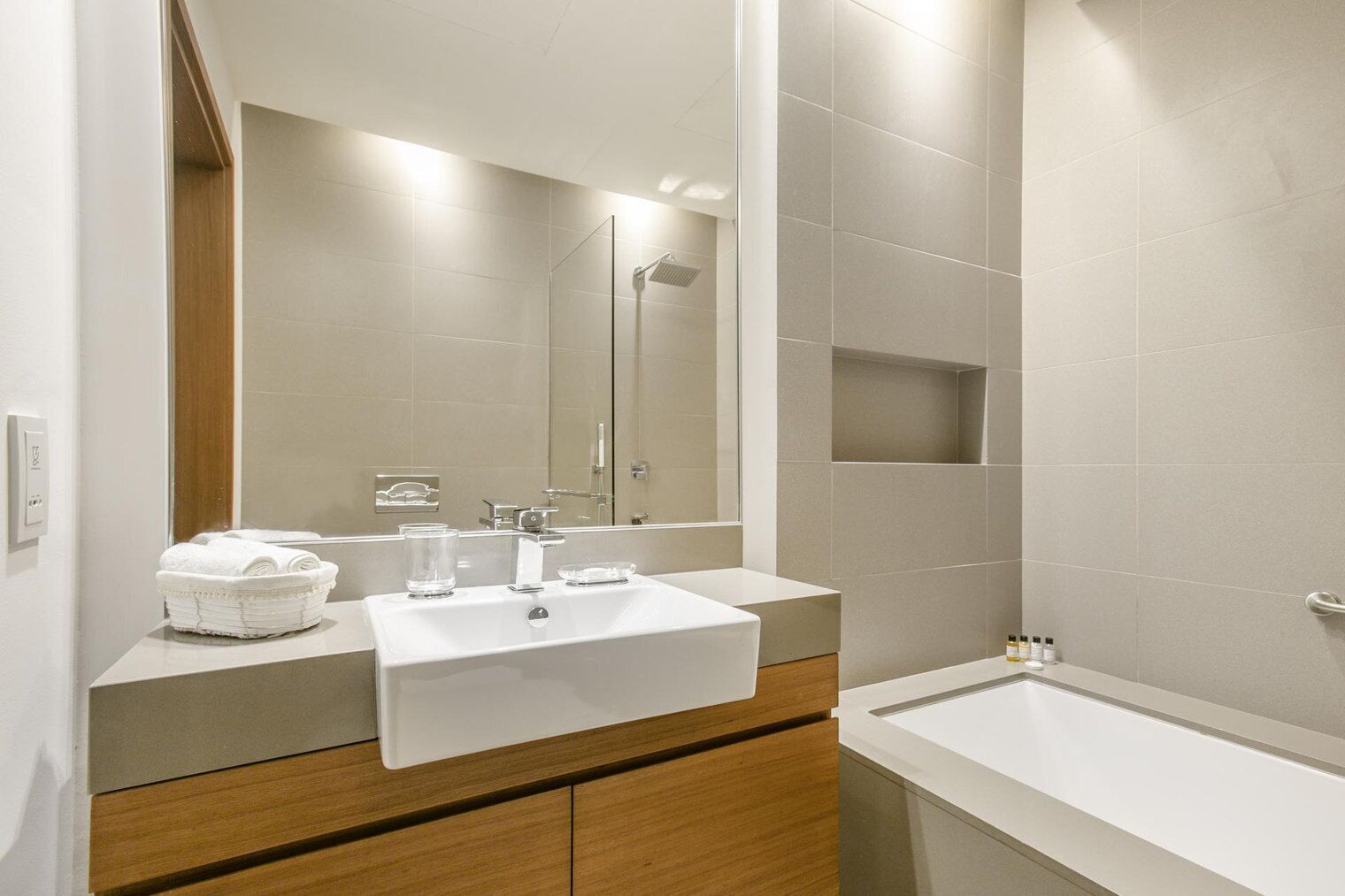 Een moderne badkamer met een witte wastafel, spiegel en badkuip. Er is een houten opbergmeubel en een handdoekhouder met opgevouwen handdoeken.