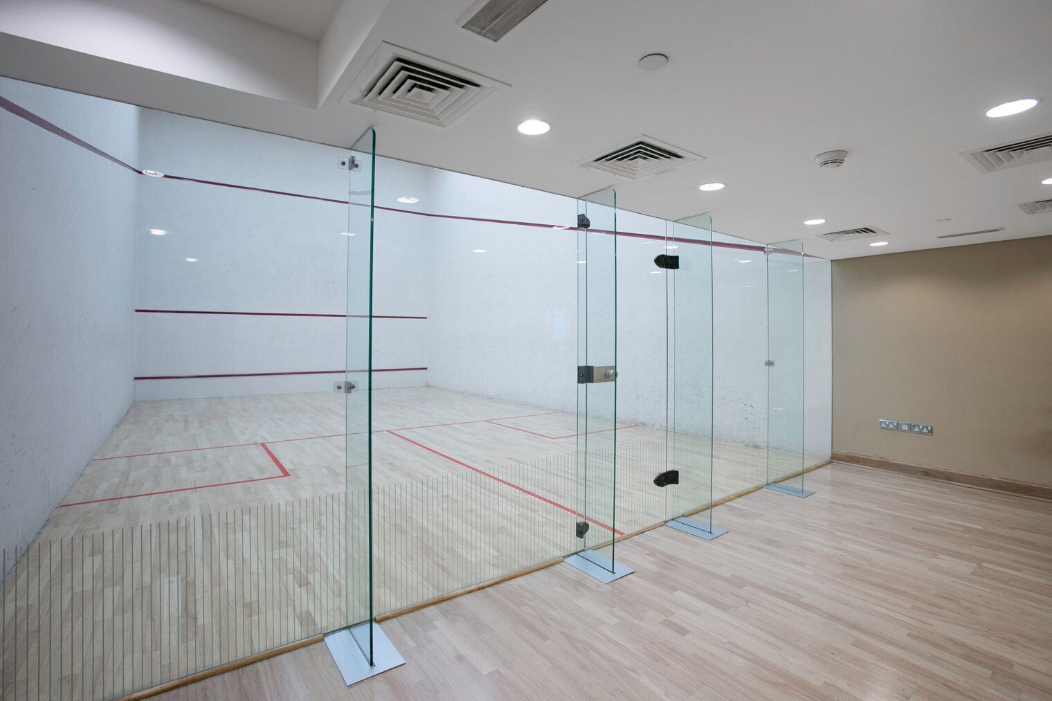 A squash court with glass walls, wooden flooring, and ceiling vents.